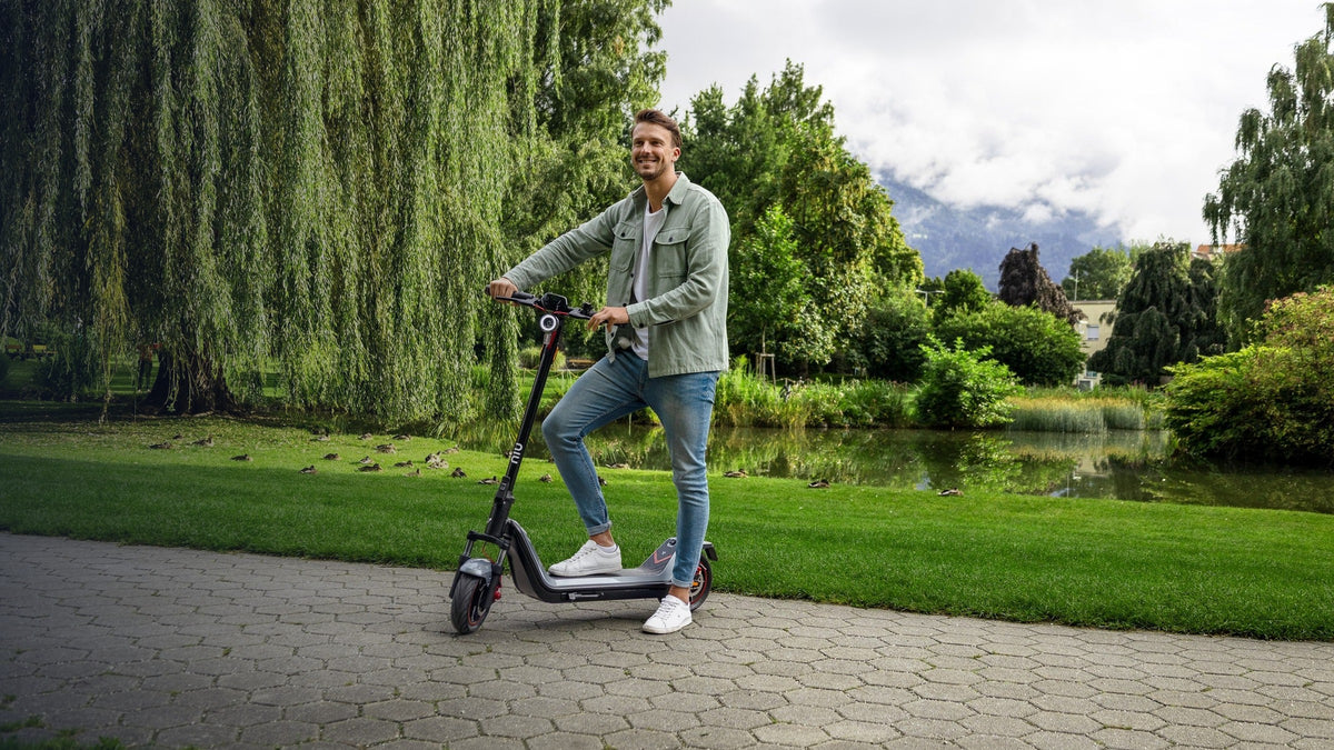 Hombre sonriendo de pie en un scooter eléctrico NIU en un parque - E-Mobility Chile, Las Condes