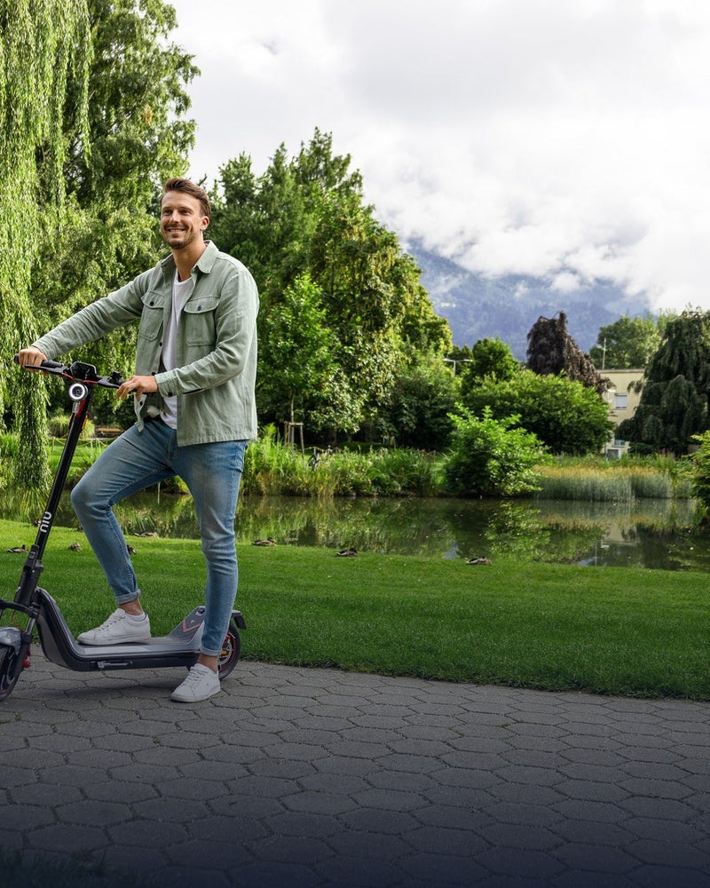 Hombre sonriendo de pie en un scooter eléctrico NIU en un parque - E-Mobility Chile, Las Condes